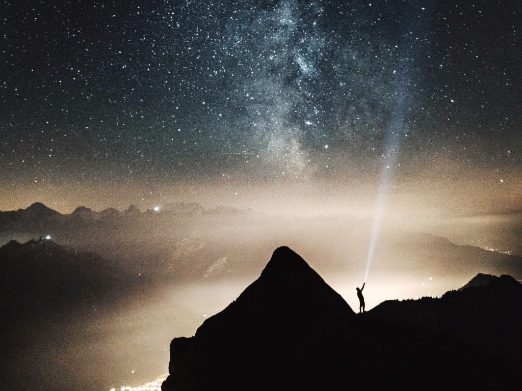 A person on a mountain peak shining a light beam into a starry night sky
