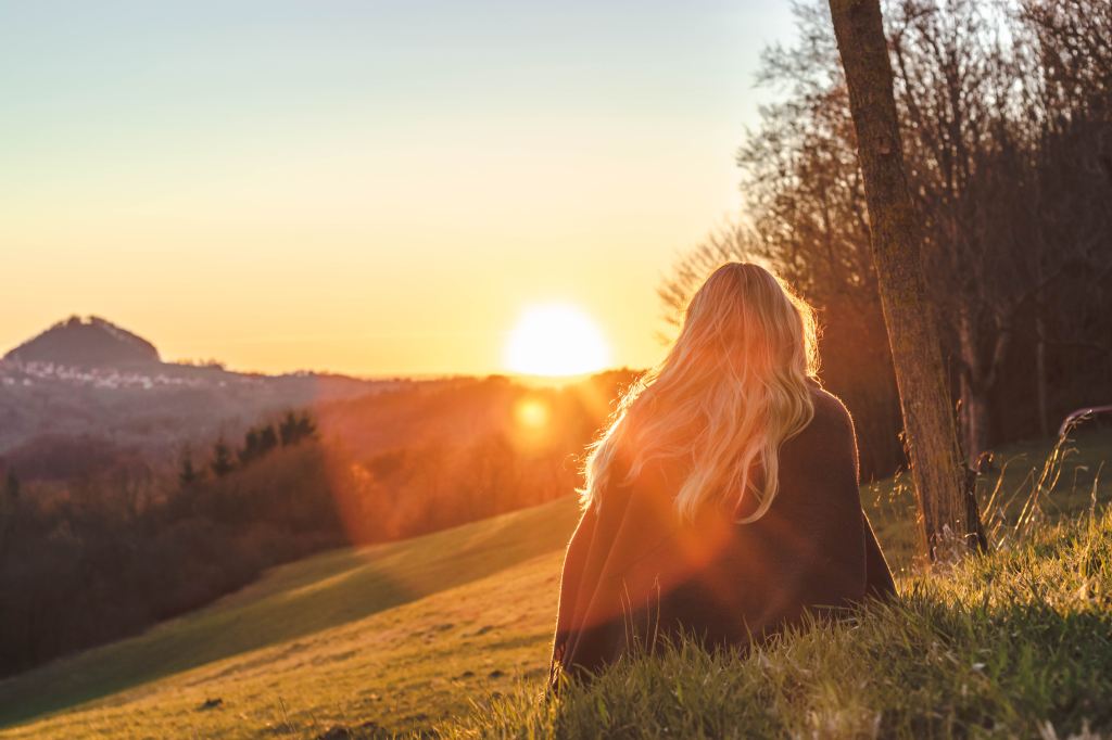 A woman with long blond hair sitting on a hillside watching the sunset