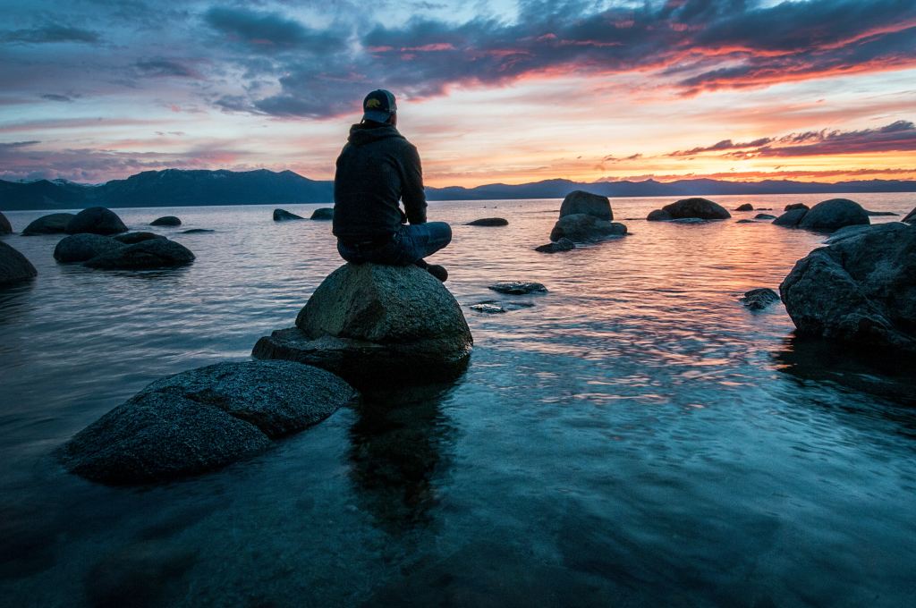 A person sitting on a rock in a lake under a colorful sunset sky