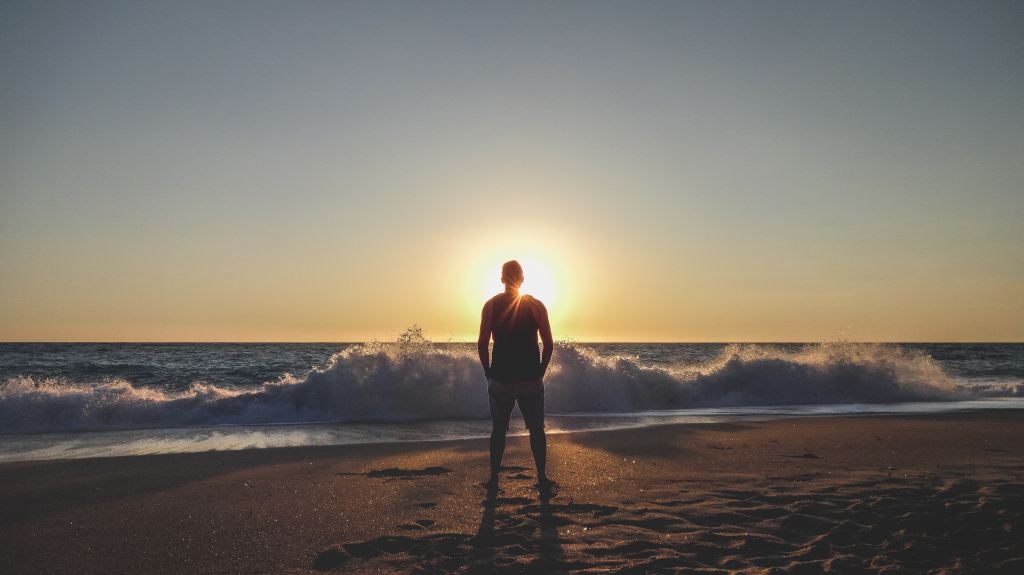 A man standing on a beach facing the sunset as waves break behind him