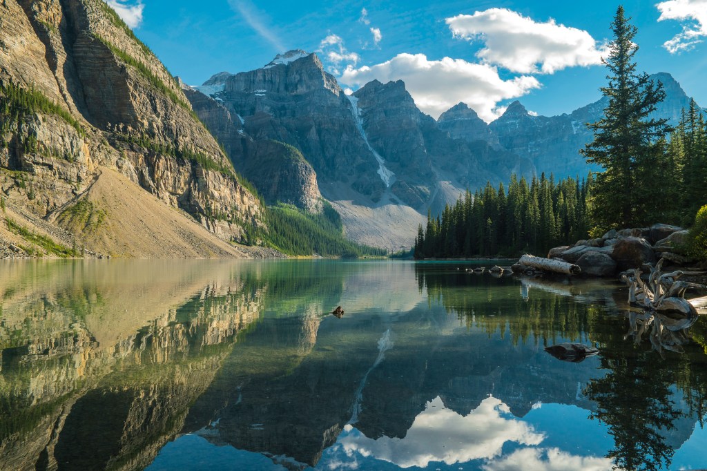 A calm mountain lake reflecting rocky peaks, pine trees, and clouds